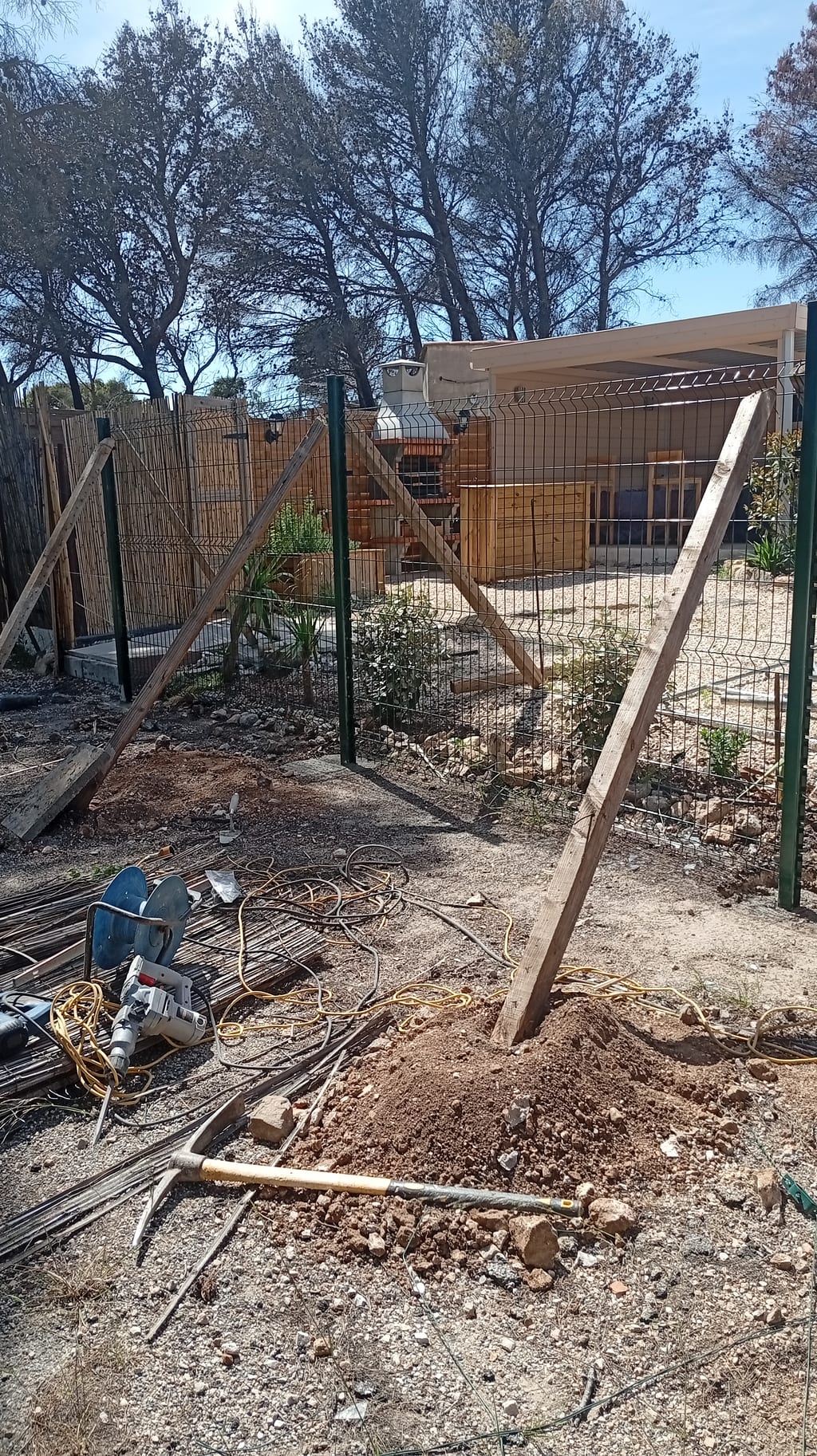 Poteau d'angle de grillage rigide étayé pendant la prise du plot béton, brise-vue bambou en arrière-plan — Les Cadeneaux